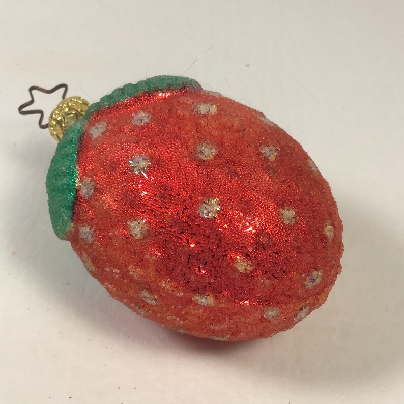 May include: A red and green glass Christmas bauble shaped like a strawberry. The strawberry has a glittery finish and white dots.