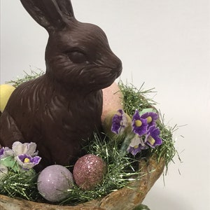 May include: A chocolate bunny sits in a nest of green and gold tinsel with purple flowers and pastel colored eggs.