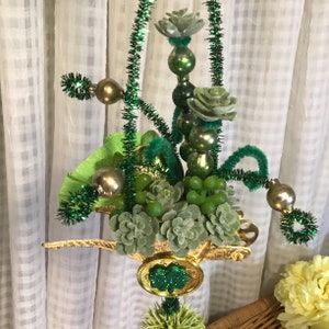 May include: A gold-colored wheelbarrow filled with green succulents, green grapes, and green and gold ornaments. The wheelbarrow is decorated with green tinsel and green and gold beads.