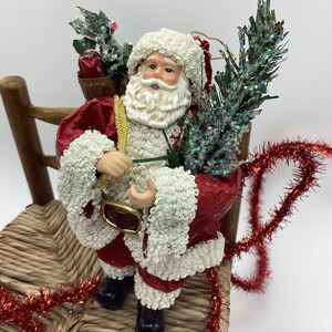 May include: A red and white Santa Claus figurine holding a small green tree and a bag of gifts. He is sitting on a wooden chair with red tinsel around it.