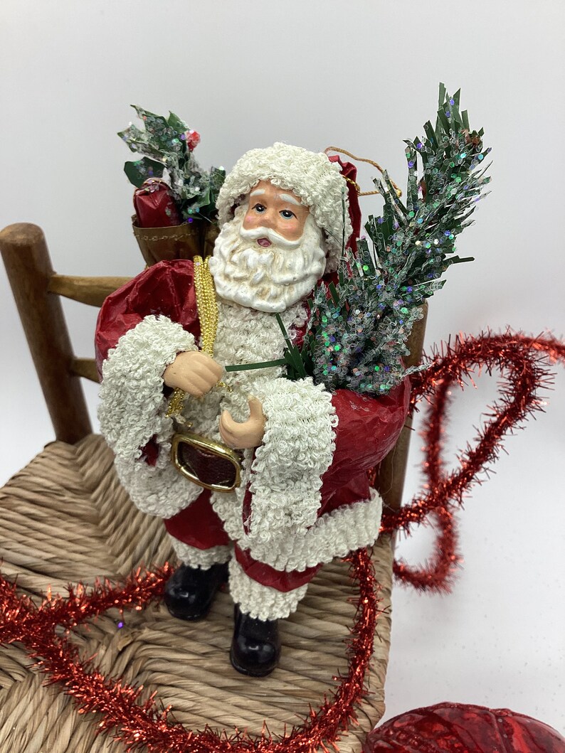 May include: A red and white Santa Claus figurine holding a small green tree and a bag of gifts. He is sitting on a wooden chair with red tinsel around it.