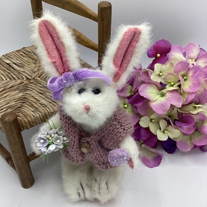 May include: A white plush bunny wearing a purple headband, a pink sweater, and holding a bouquet of purple flowers and a purple egg. The bunny is sitting on a wooden chair with a woven seat.