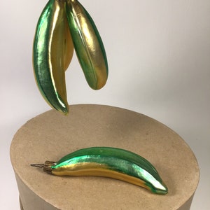 May include: Three banana-shaped Christmas ornaments in green and gold. The ornaments have a shiny, reflective surface. One ornament rests on a brown surface, while two others hang from a wire. The ornaments have a vintage appearance.