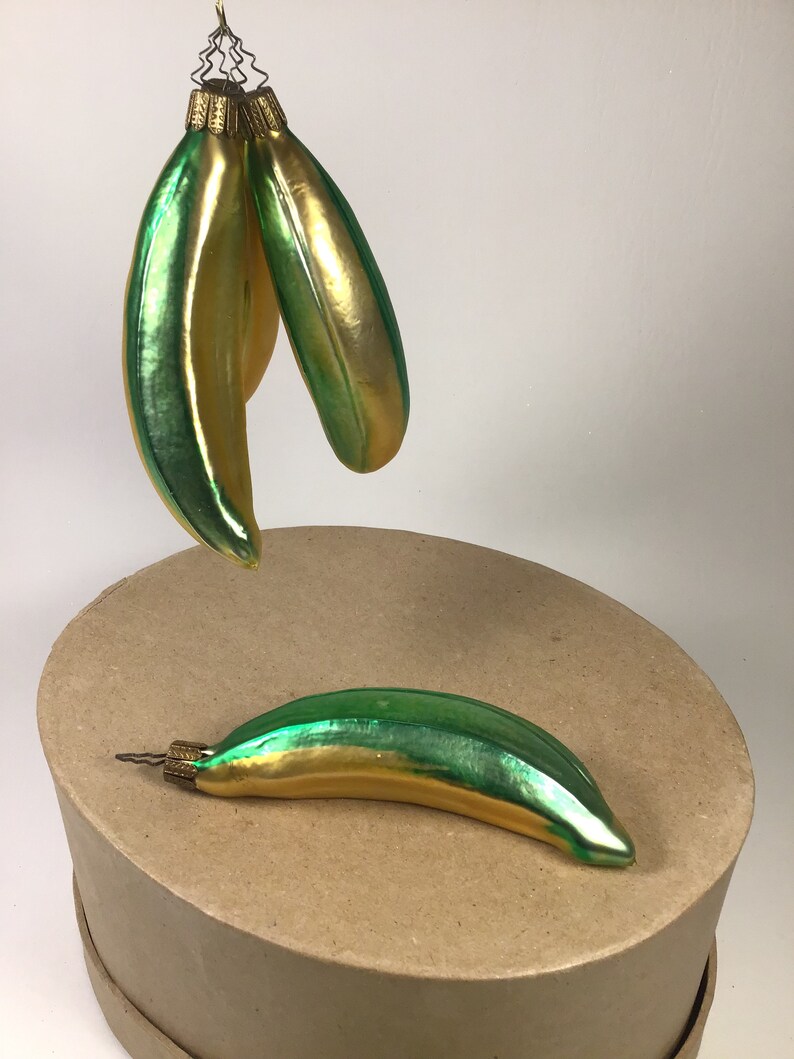 May include: Three banana-shaped Christmas ornaments in green and gold. The ornaments have a shiny, reflective surface. One ornament rests on a brown surface, while two others hang from a wire. The ornaments have a vintage appearance.