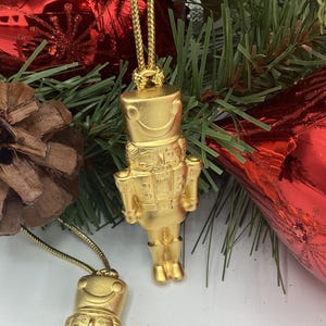 Puede incluir: Adornos navideños dorados con forma de cascanueces colgando de una exhibición festiva verde y roja. Los adornos están detallados con una cara sonriente y un uniforme. El fondo incluye agujas de pino, una piña y adornos rojos.