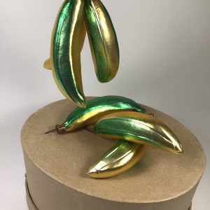 May include: Banana-shaped Christmas decorations with a gold and green metallic finish. The ornaments are suspended from a wire and are on a round, cardboard box. The decorations have a vintage appearance.
