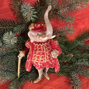 May include: A red and gold Christmas ornament of an elephant wearing a red and gold hat and a red and gold coat. The elephant is holding a gold cane.