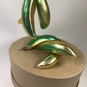 May include: Glass banana Christmas ornaments in gold and green. The ornaments are shaped like bananas and have a shiny, reflective surface. The ornaments are on a brown cardboard box.