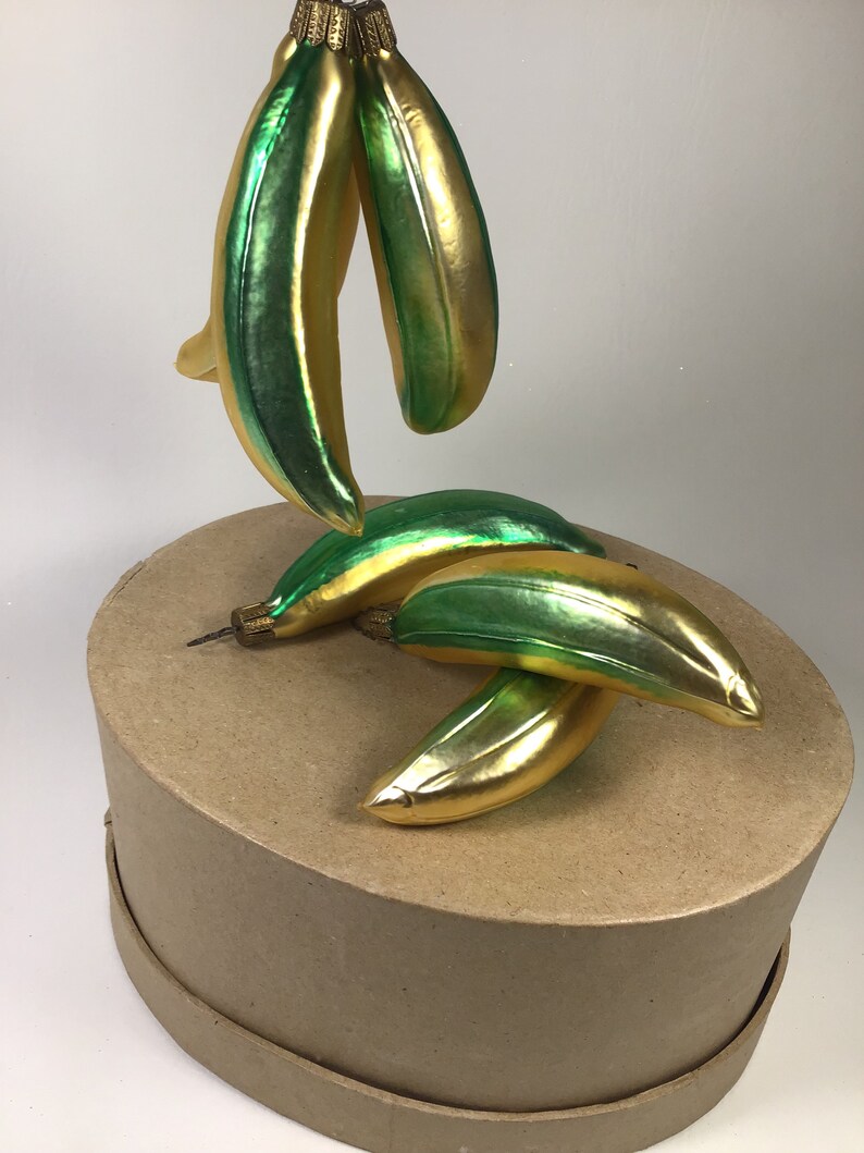 May include: Glass banana Christmas ornaments in gold and green. The ornaments are shaped like bananas and have a shiny, reflective surface. The ornaments are on a brown cardboard box.