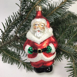 May include: A festive Christmas ornament shaped like Santa Claus, featuring a red coat, white trim, and a black belt. The ornament has green mittens and is set against a backdrop of green pine needles.