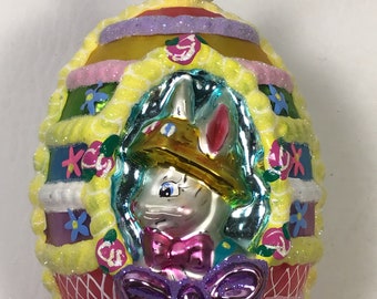 以下が含まれることがあります： 白いウサギがピンクのリボンと金の帽子をかぶったガラス製のイースターエッグのオーナメント。卵は黄色、ピンク、青、緑のフロスティングとスプリンクルで飾られています。