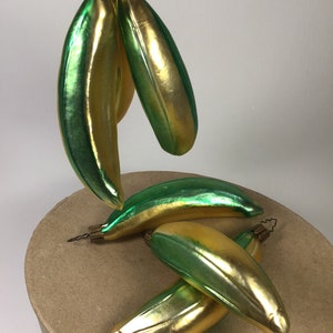 May include: A collection of banana-shaped Christmas tree decorations. The baubles are a blend of gold and green, with a glossy finish. Each has a gold cap and a wire loop for hanging. The ornaments are arranged on a round, cardboard surface.