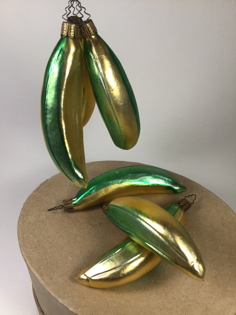 May include: A collection of banana-shaped Christmas tree decorations. The baubles are a blend of gold and green, with a glossy finish. Each has a gold cap and a wire loop for hanging. The ornaments are arranged on a round, cardboard surface.