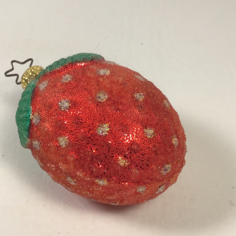 May include: A red and green glass Christmas bauble shaped like a strawberry. The strawberry has a textured surface and white glitter accents.