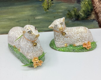 May include: Two white ceramic lambs with gold accents, sitting on a bed of green grass with small orange flowers. Each lamb has a gold bell around its neck.