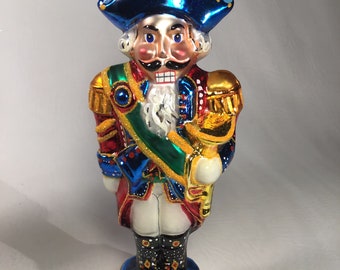 May include: A colorful glass nutcracker ornament with a blue hat, red and white uniform, and a gold trumpet. The ornament is decorated with glitter and has a gold base.