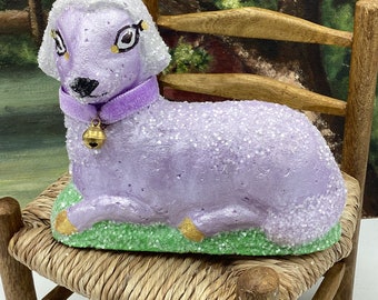 May include: A purple, sparkly, lamb figurine with a white fluffy head, a purple velvet collar, and a gold bell. The lamb is sitting on a green grass base and is on a wooden chair.
