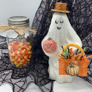 Puede incluir: Una figurita de fantasma blanco con un sombrero marrón, sosteniendo una calabaza y una bolsa de maíz dulce. El fantasma está de pie sobre una tela de encaje negro con un frasco de maíz dulce en el fondo.