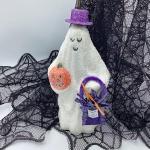 May include: A white ghost figurine with a purple hat and a pumpkin in its hand. The ghost is holding a purple trick or treat bag with orange and white candy corn. The ghost is standing on a black lace background.