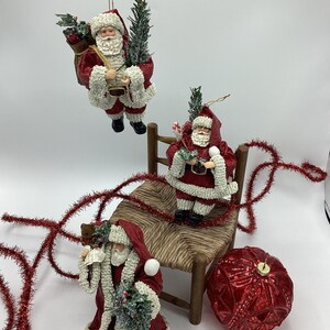 May include: Three red and white Santa Claus ornaments, one sitting on a wooden chair, one holding a small tree, and one holding a bag of gifts. The ornaments are decorated with white beads and glitter. A red and gold Christmas bauble is also in the image.