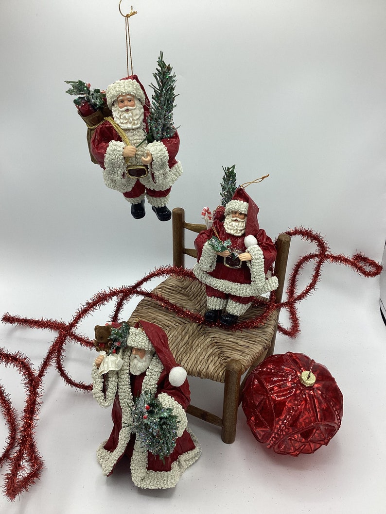 May include: Three red and white Santa Claus ornaments, one sitting on a wooden chair, one holding a small tree, and one holding a bag of gifts. The ornaments are decorated with white beads and glitter. A red and gold Christmas bauble is also in the image.