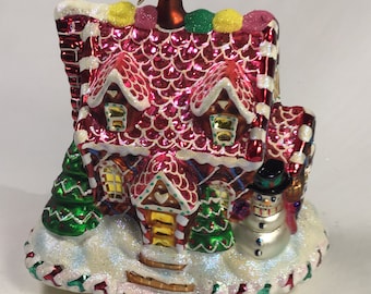 May include: A red and white gingerbread house Christmas ornament with a snowman standing in front of it. The house has candy cane trim and a white frosted roof. The ornament is covered in glitter.