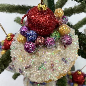 May include: A white, glittery Christmas bauble with a red, glittery teapot on top. The teapot is surrounded by colourful, glittery balls.