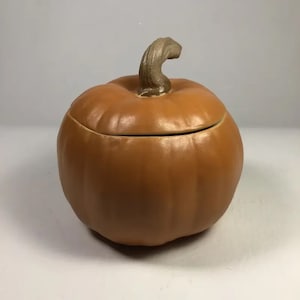 Pode incluir: Um recipiente de cerâmica em forma de abóbora com uma tampa. O recipiente é de cor castanho claro e tem uma superfície ligeiramente texturizada. A tampa tem a forma de um caule de abóbora.