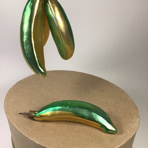 May include: Glass banana Christmas decorations with a green and gold metallic finish. One ornament rests on a round cardboard surface, while two others hang above. The decorations have a vintage style and are ready to hang on a Christmas tree.