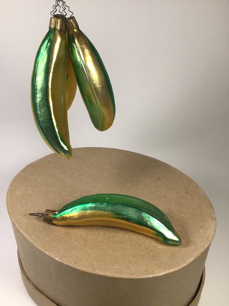 May include: Glass banana Christmas decorations with a green and gold metallic finish. One ornament rests on a round cardboard surface, while two others hang above. The decorations have a vintage style and are ready to hang on a Christmas tree.