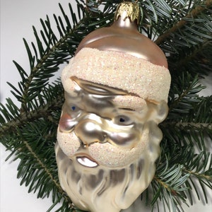May include: A vintage glass Christmas bauble shaped like Santa Claus's head. The bauble is a pale gold colour with a white glitter beard and hat. The bauble has blue eyes and a pink nose.