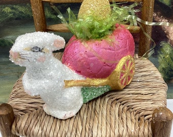 May include: A white bunny figurine pulling a pink wagon with a gold glitter egg inside. The wagon is decorated with green and gold accents. The bunny and wagon are sitting on a woven chair.