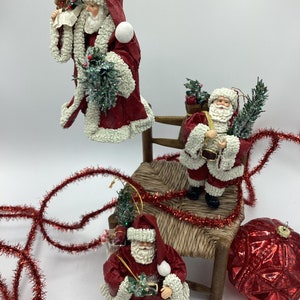 May include: Three red and white paper mache Santa Claus ornaments with green holly and berries. The ornaments are hanging against a white background. The Santa Claus on the left is holding a teddy bear and a piece of paper. The Santa Claus in the middle is holding a sack and a sprig of holly. The Santa Claus on the right is holding a small gift box and a sprig of holly.