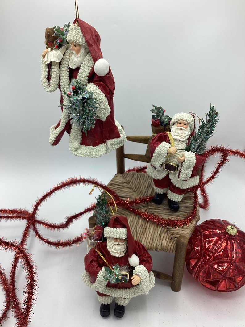 May include: Three red and white paper mache Santa Claus ornaments with green holly and berries. The ornaments are hanging against a white background. The Santa Claus on the left is holding a teddy bear and a piece of paper. The Santa Claus in the middle is holding a sack and a sprig of holly. The Santa Claus on the right is holding a small gift box and a sprig of holly.