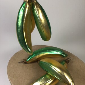 May include: A collection of banana-shaped Christmas tree decorations. The baubles are a blend of gold and green, with a glossy finish. Each ornament has a gold-coloured metal cap and a wire loop for hanging. The decorations are arranged on a neutral surface.