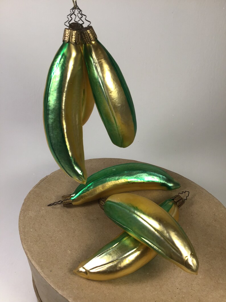 May include: A collection of banana-shaped Christmas tree decorations. The baubles are a blend of gold and green, with a glossy finish. Each ornament has a gold-coloured metal cap and a wire loop for hanging. The decorations are arranged on a neutral surface.