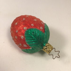 May include: A red and green glass Christmas bauble shaped like a strawberry with white glitter accents. The bauble has a gold star-shaped hanger.