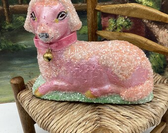 May include: A pink lamb figurine with a sparkly finish and a pink velvet collar with a gold bell. The lamb is sitting on a small wooden chair with a woven seat.
