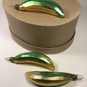 May include: Three vintage Christmas tree decorations shaped like bananas. The ornaments are green and gold with a metallic finish. Each has a small metal cap with a wire loop. One ornament rests on a brown box.