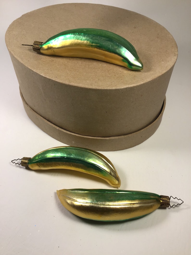 May include: Three vintage Christmas tree decorations shaped like bananas. The ornaments are green and gold with a metallic finish. Each has a small metal cap with a wire loop. One ornament rests on a brown box.