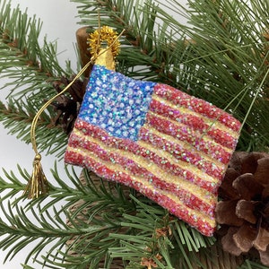 May include: A glass ornament shaped like an American flag with red and white stripes and blue stars. The ornament is covered in glitter and has a gold tassel.