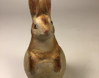 May include: A ceramic rabbit figurine sitting on a cracked egg. The rabbit is brown with white fur and has painted brown eyes. The egg is a light brown color.