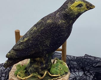 May include: A black and gold glitter raven figurine perched on a small wooden chair with moss and a gold trim. The raven has a yellow beak and eyes.