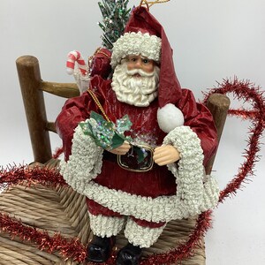 May include: A red paper mache Santa Claus ornament sitting on a wicker chair. He is holding a small green sprig and has a white beard and white fur trim on his red suit. He is carrying a small Christmas tree and a candy cane.
