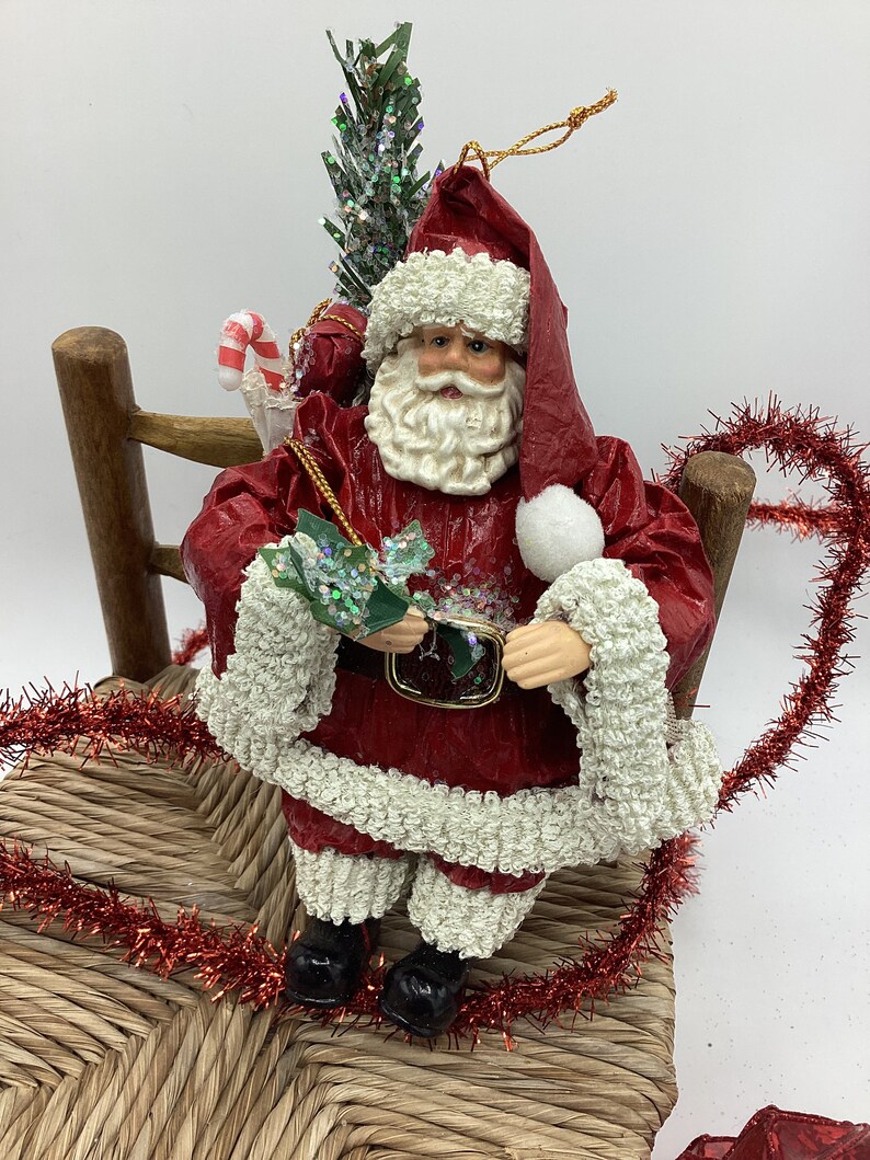 May include: A red paper mache Santa Claus ornament sitting on a wicker chair. He is holding a small green sprig and has a white beard and white fur trim on his red suit. He is carrying a small Christmas tree and a candy cane.