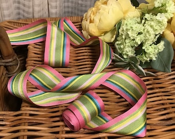 May include: A length of striped ribbon in pink, green, yellow, and blue, displayed on a wicker basket. The ribbon is likely for crafts or gift wrapping. Yellow flowers and foliage are in the background.