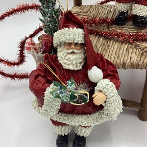 May include: A red and white Santa Claus ornament with a white beard and a green holly sprig in his hand. He is wearing a red suit with white trim and black boots. He is holding a small gift box in his other hand.