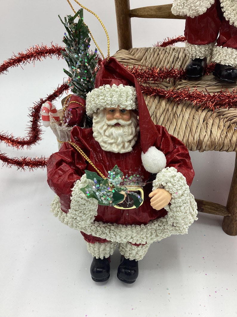 May include: A red and white Santa Claus ornament with a white beard and a green holly sprig in his hand. He is wearing a red suit with white trim and black boots. He is holding a small gift box in his other hand.
