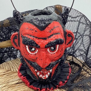 May include: A red and black devil head Halloween decoration with a black ruffled collar. The devil has a black beard, white teeth, and black horns.