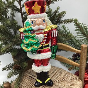 May include: A Christmas ornament shaped like a nutcracker soldier. The ornament is red, gold, white, and green, with a crown and a small Christmas tree. The nutcracker is holding a green Christmas tree. The ornament is sitting on a small chair.